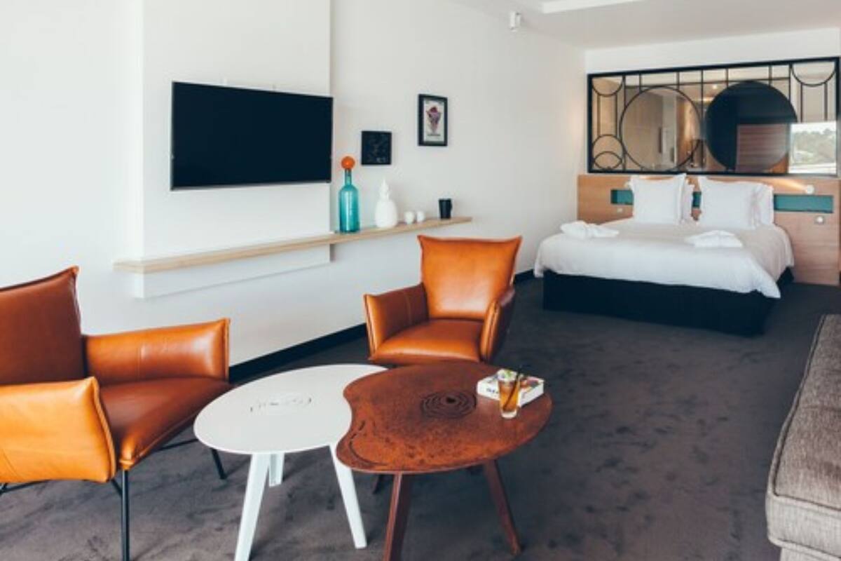A hotel room with a bed, two leather armchairs, a small round table, a TV mounted on the wall, and a window with a view.