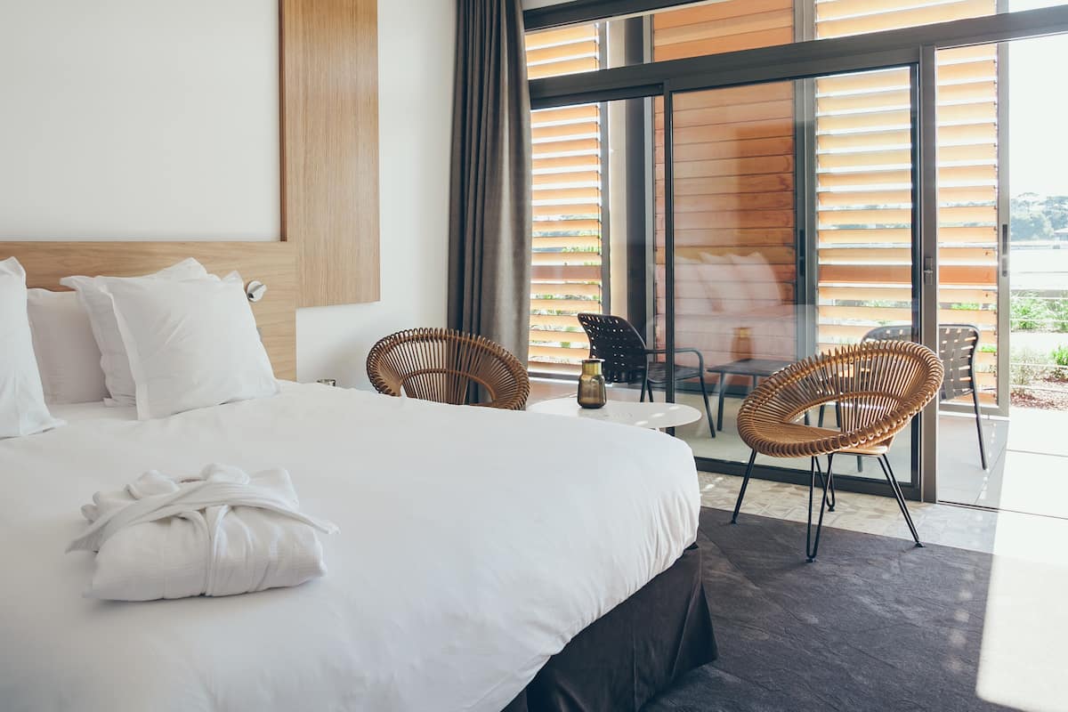A modern hotel room with a large bed, two wicker chairs, a small table, and a view of the outdoors through large windows.