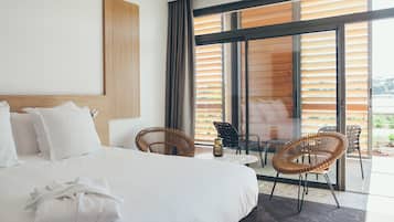 Ein modernes Hotelzimmer mit einem großen Bett, zwei Korbstühlen, einem kleinen Tisch und Blick ins Freie durch große Fenster.