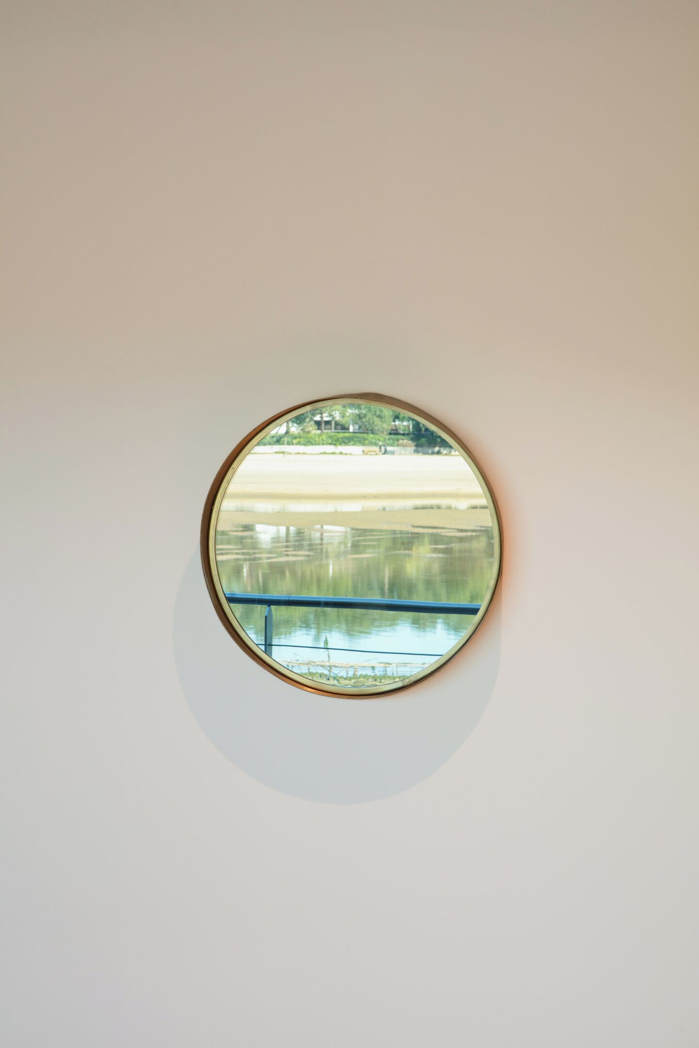 A round mirror on a wall reflecting a calm body of water and a bridge.