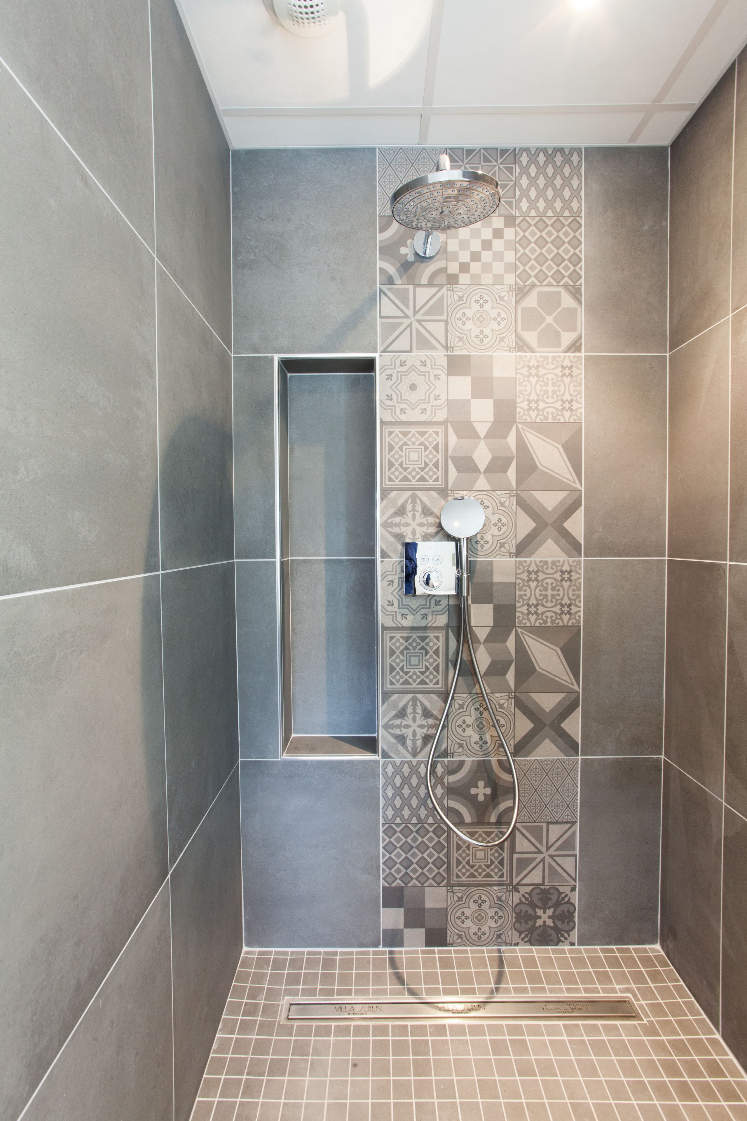 Ducha con cabezal de ducha manual y grifo empotrado en la pared. La ducha tiene suelo y paredes de azulejos con diseños geométricos.