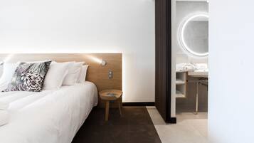 Une chambre d’hôtel moderne avec un lit bien fait, une petite table d’appoint en bois et une salle de bain dotée d’un miroir rond et d’un lavabo.