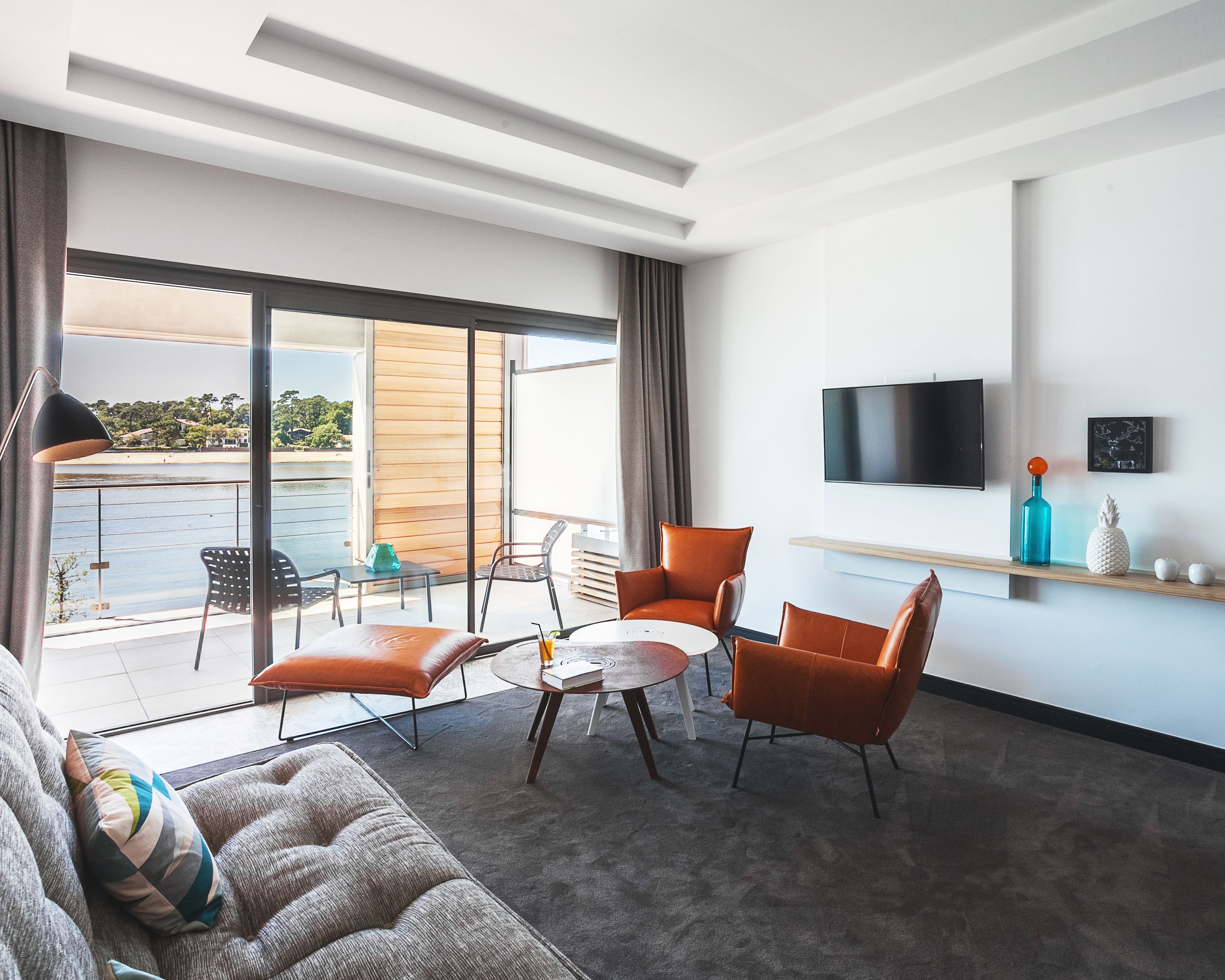 A modern living room with a sofa, two armchairs, a coffee table, and a TV mounted on the wall. There is a balcony with a view of the water and trees.