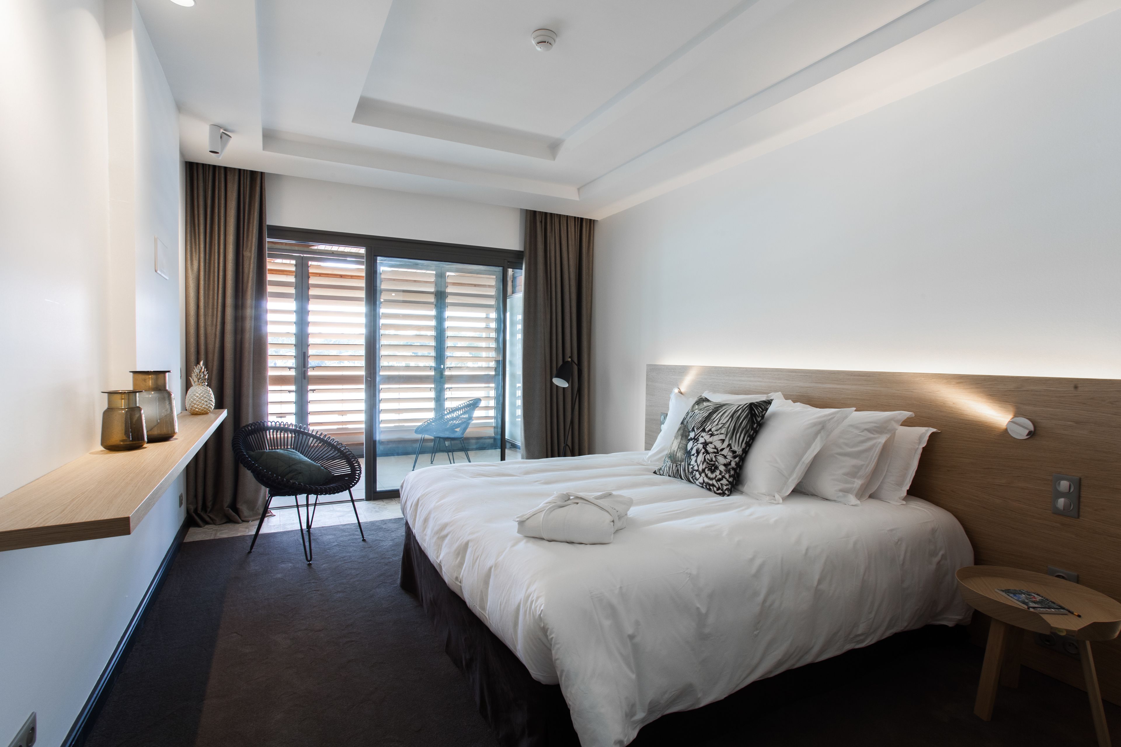 A modern hotel room with a large bed, a small desk, a chair, and a view of the outside through a sliding door.