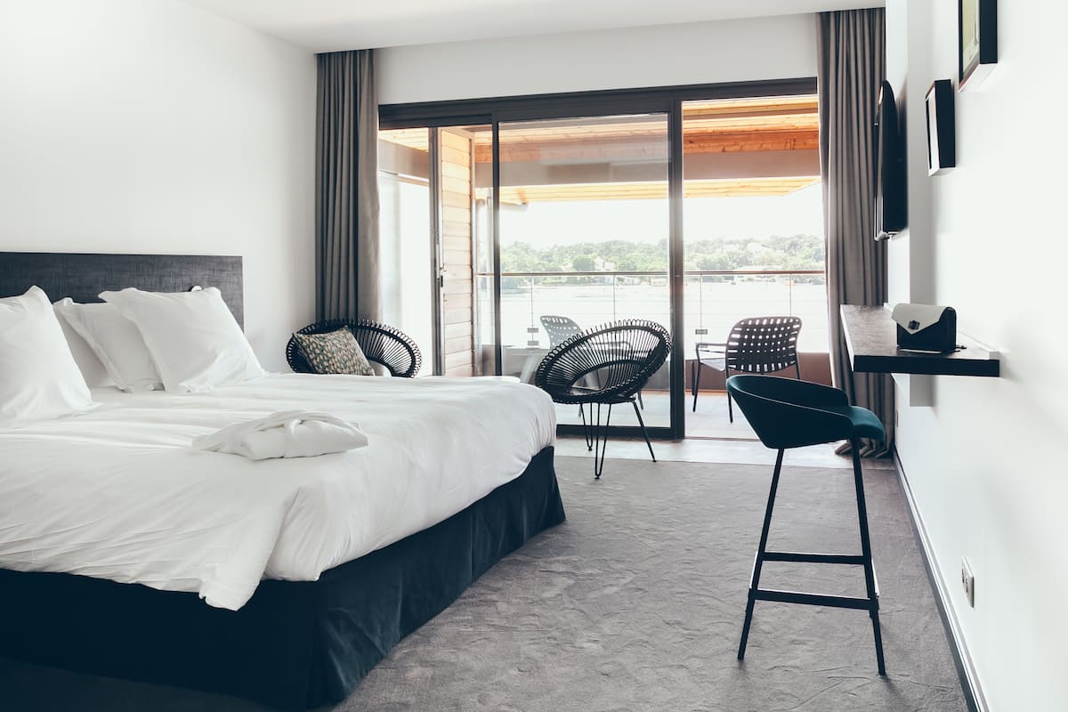 A modern hotel room with a large bed, a balcony with chairs, and a small table.