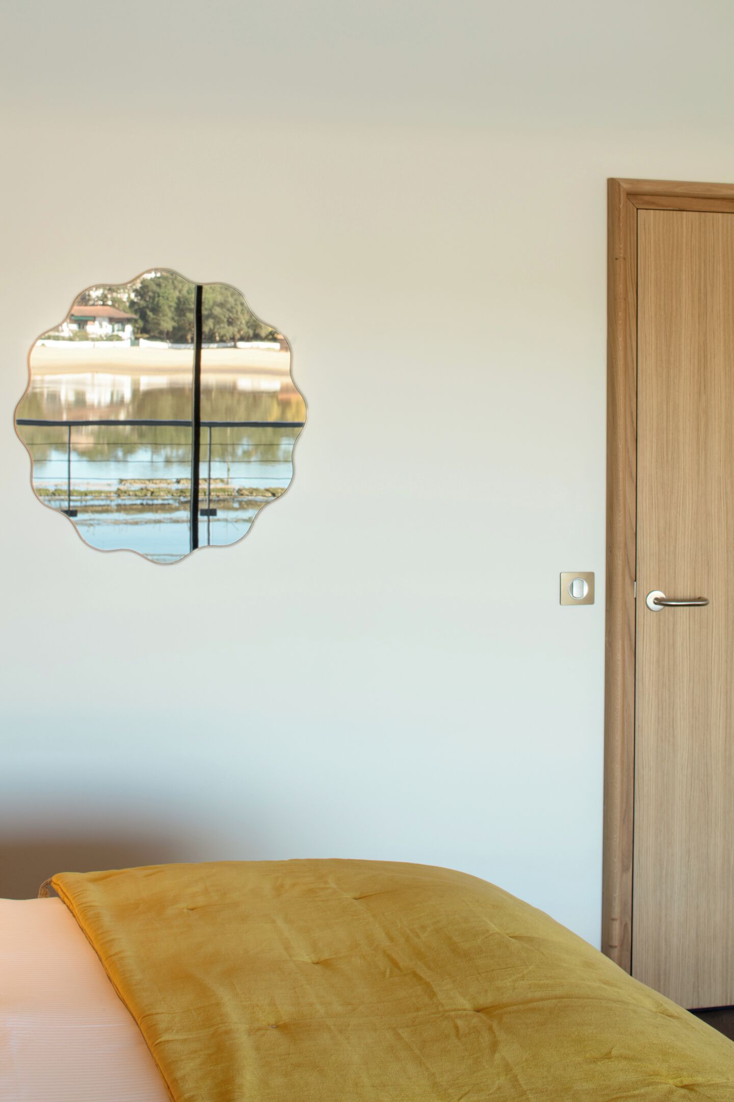 Ein Zimmer mit einem Bett, einer Holztür und einem Wandspiegel, der ein Gebäude und Bäume reflektiert.