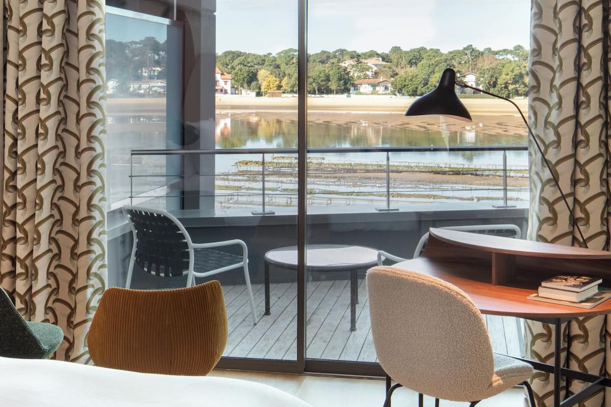 A hotel room with a large bed, a desk, and a chair. There is a balcony with a view of a lake and hills.