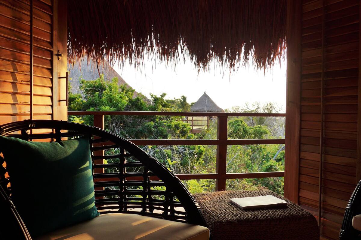 A wooden room with a thatched roof, a wicker chair with a cushion, and a view of greenery and a thatched-roof structure outside.