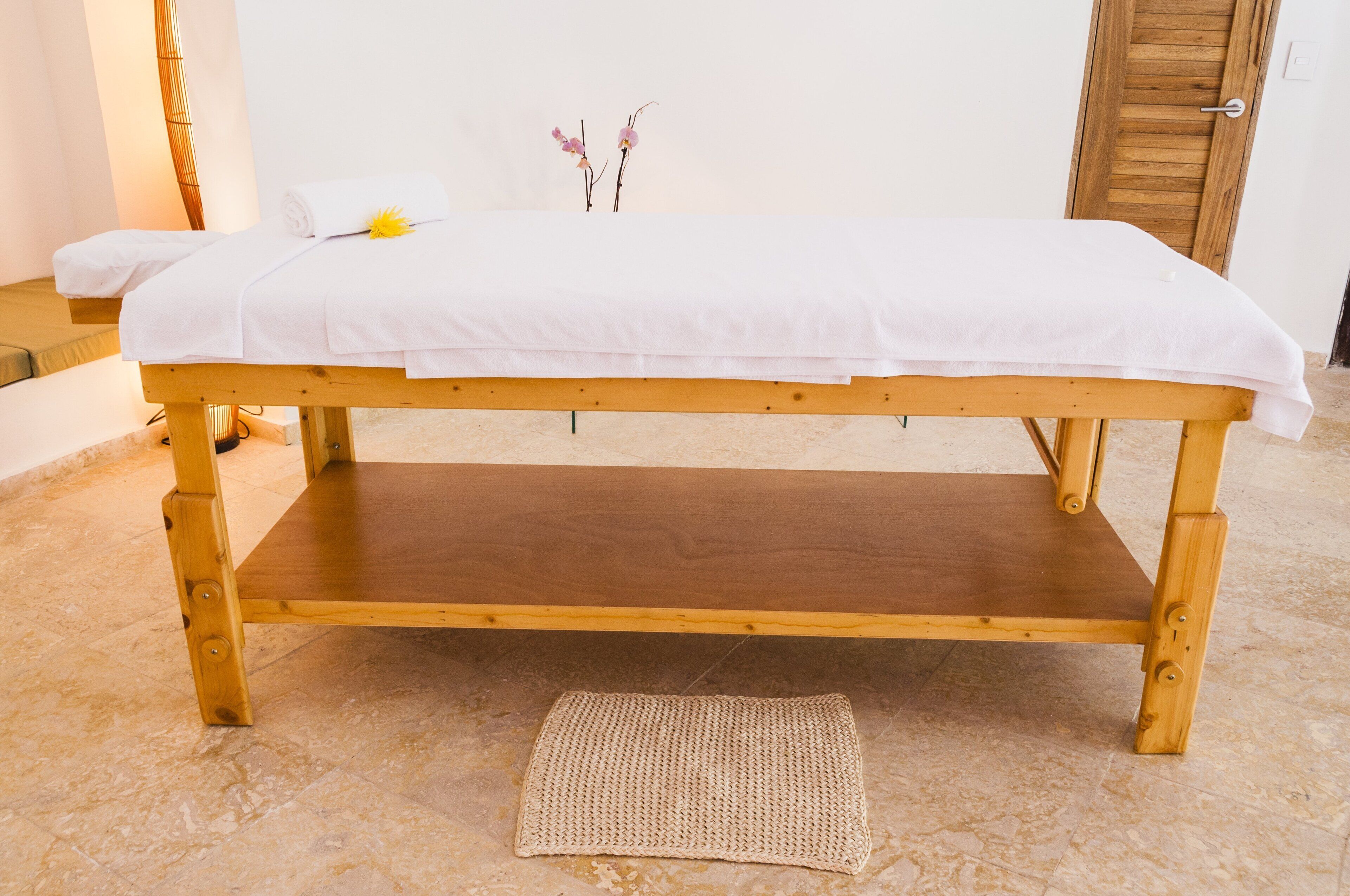 A wooden massage table with a white sheet, a woven mat, and a small flower arrangement on the wall behind.