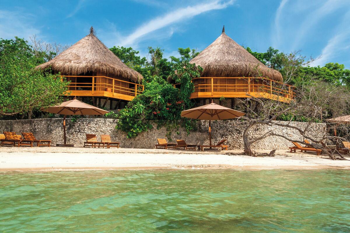 A beachside resort with thatched-roof overwater bungalows, clear turquoise water, and lounge chairs.