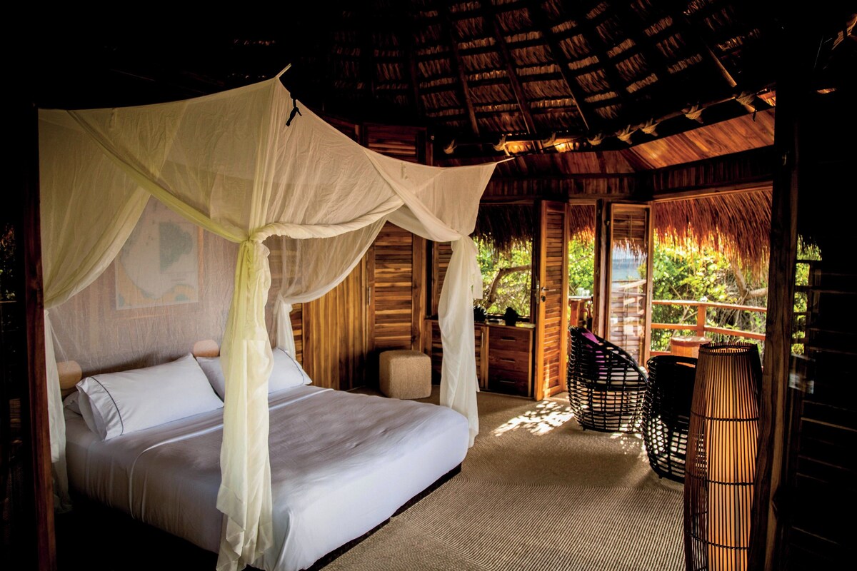 A room with a bed under a canopy, wooden walls, and a thatched roof.