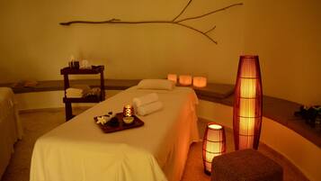 Un cuarto de spa con una camilla de masaje, toallas, velas y una rama decorativa en la pared.