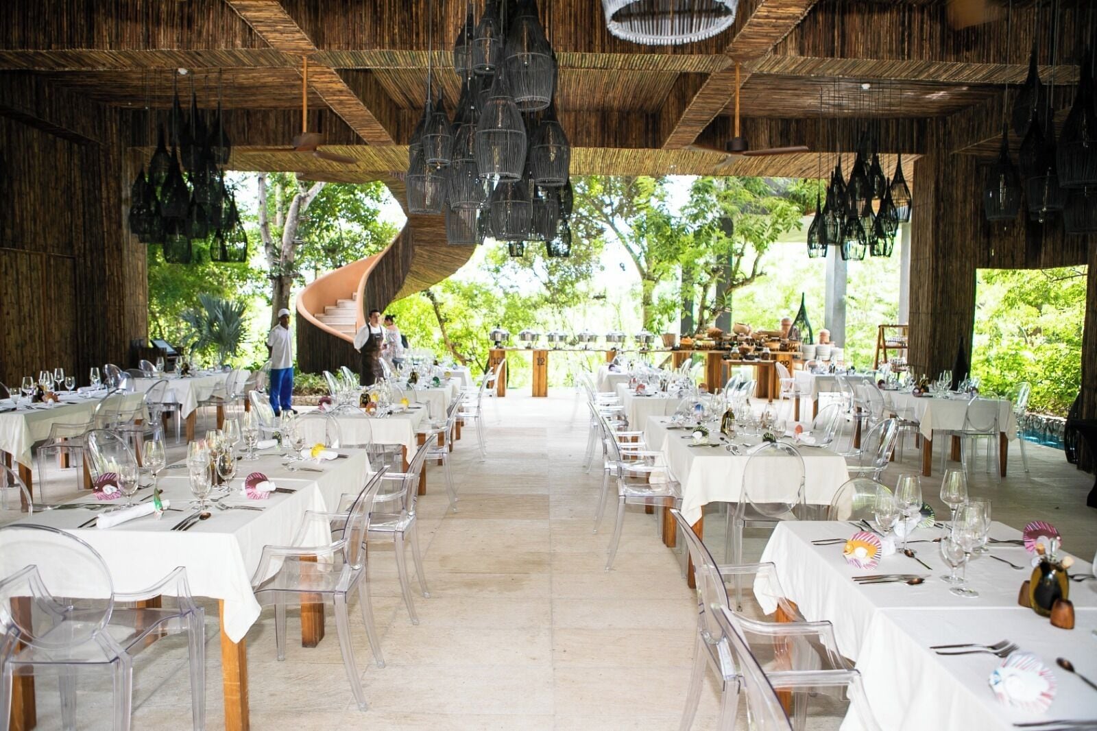A spacious outdoor dining area with multiple tables set for a meal, clear chairs, and a central spiral staircase.