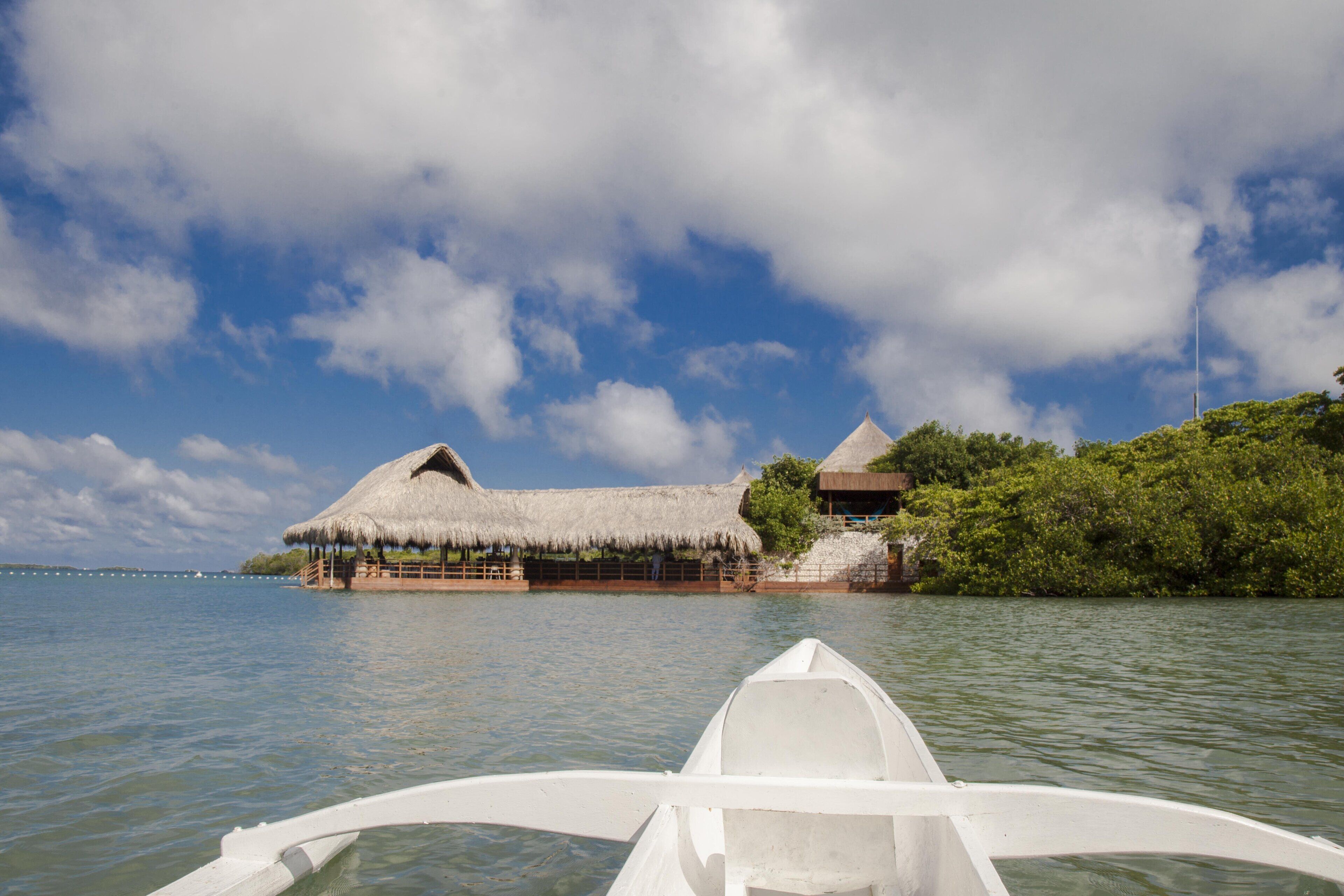 A boat with a thatched roof structure on a calm body of water, surrounded by lush greenery and a partly cloudy sky.