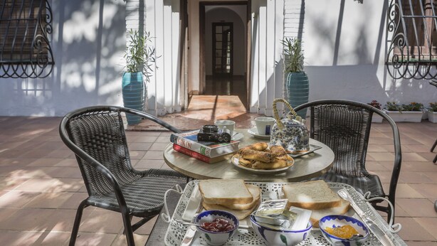 Un espacio de comedor al aire libre con sillas de mimbre, una mesa redonda y diferentes alimentos como pan, salsas y una tetera.