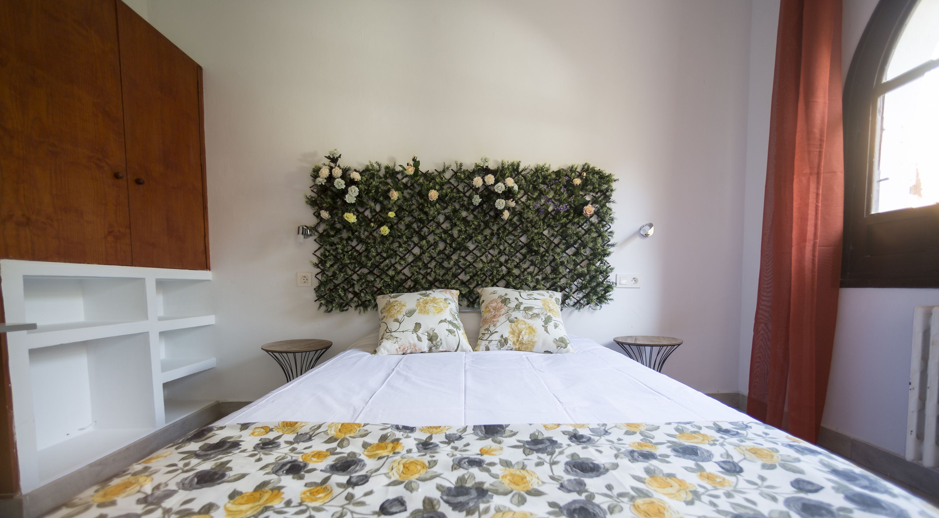 A bedroom with a bed, two nightstands, a wooden wardrobe, a wall-mounted plant, and a window with curtains.