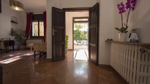 Un umbral abierto que da a un patio con vistas al exterior. Hay un radiador en la pared a la derecha y una maceta con flores moradas a la izquierda.