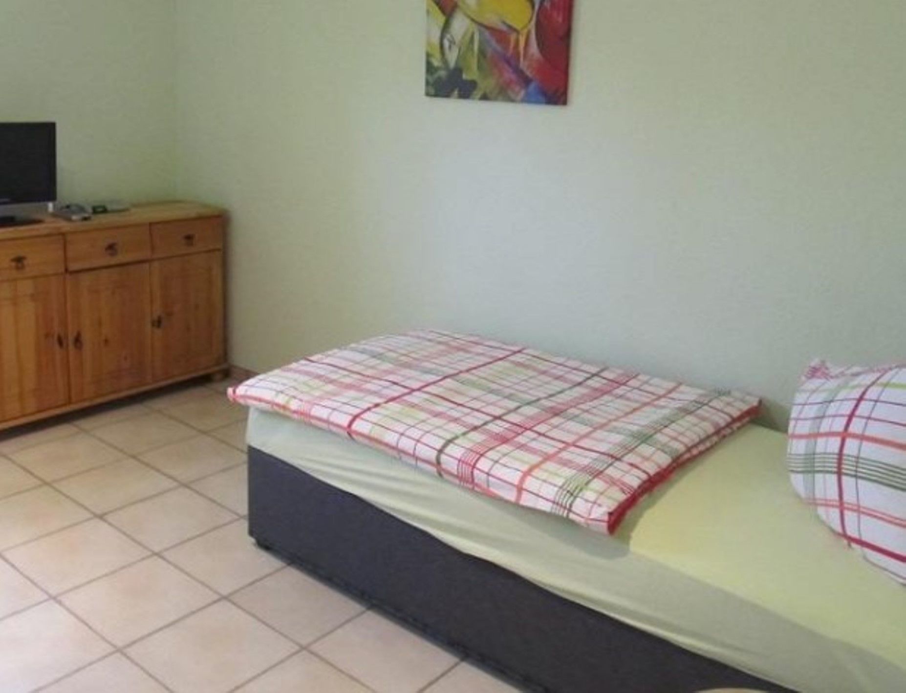 A bedroom with a bed, a wooden dresser, and a wall-mounted TV.