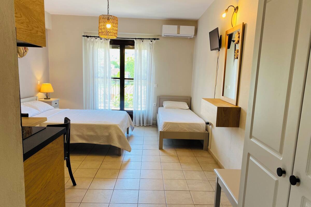 A hotel room with two beds, a desk, a chair, a lamp, a hanging light fixture, a wall-mounted TV, and a window with curtains.