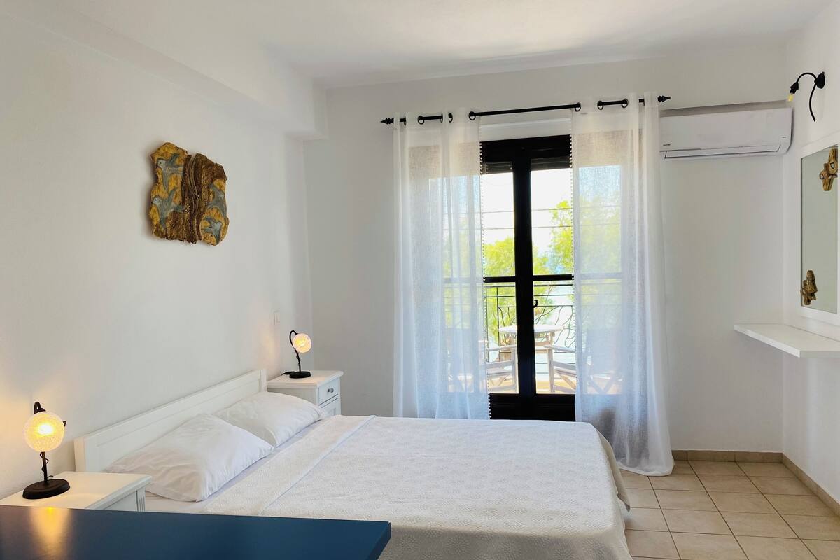 A bedroom with a bed, bedside lamps, a window with curtains, a small table, and a wall-mounted shelf.