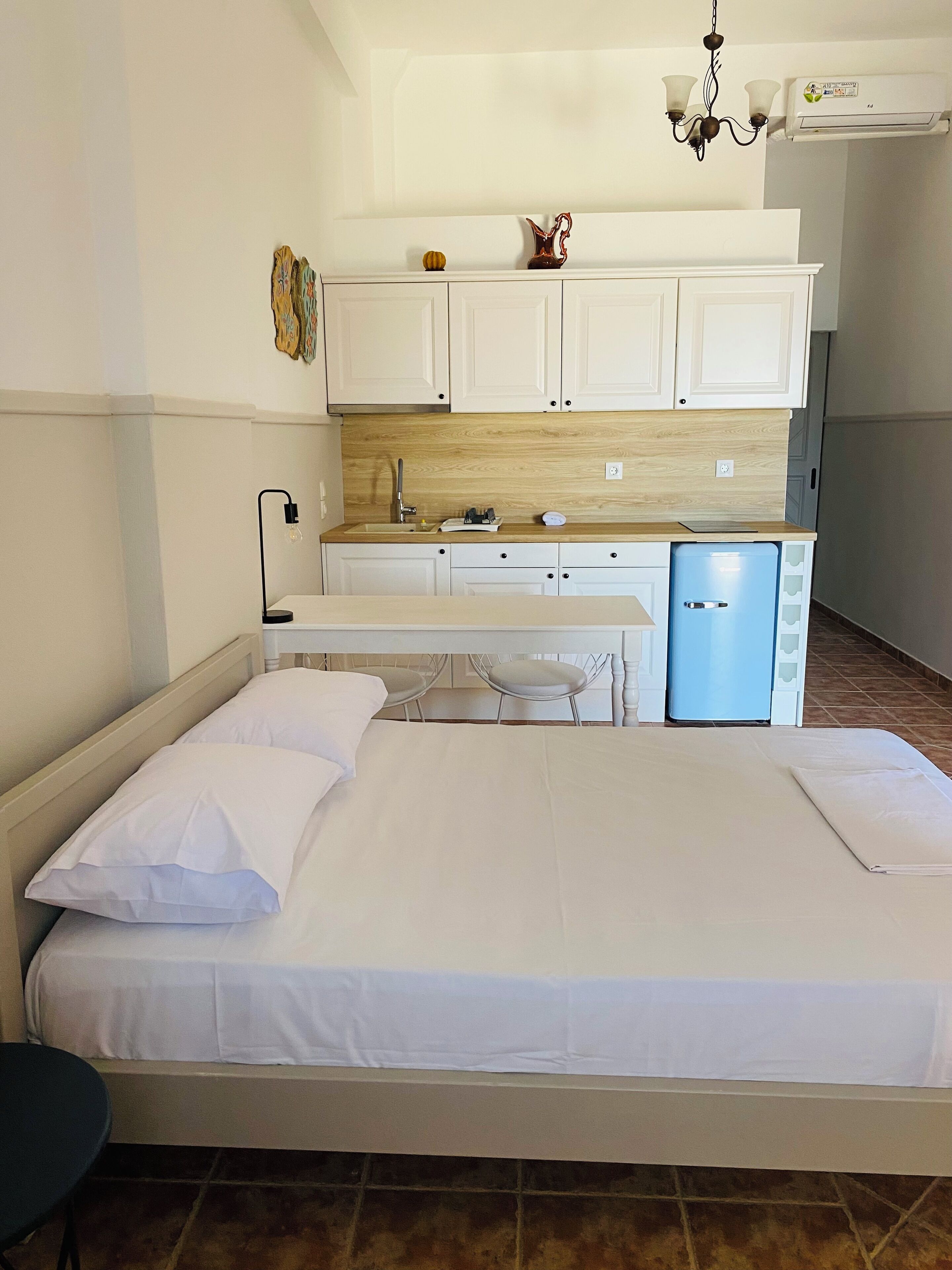 A bedroom with a bed, a small desk, and a kitchenette area with cabinets and a refrigerator.