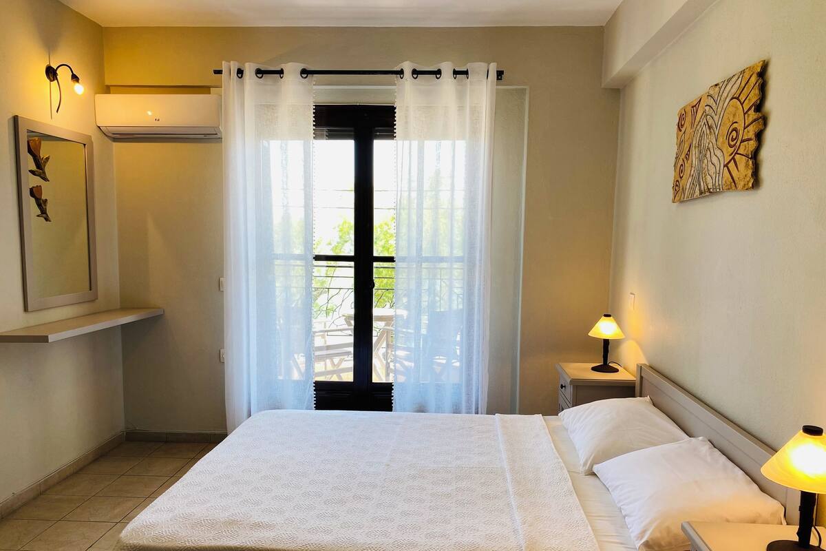 A bedroom with a bed, a nightstand, a lamp, a window with curtains, a wall-mounted artwork, and a ceiling fan.