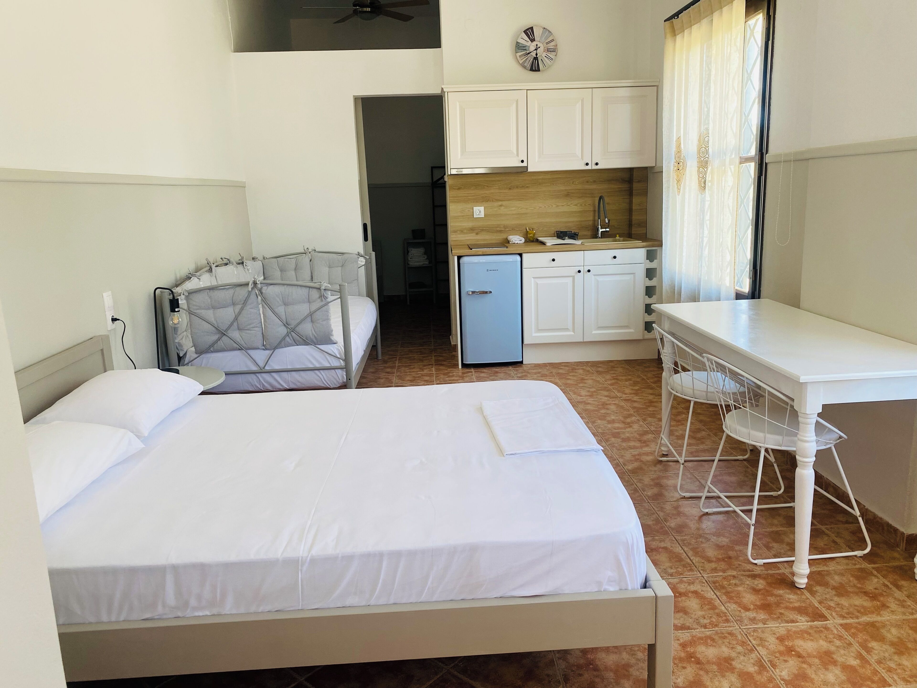 A hotel room with a bed, a desk, a chair, a kitchenette, and a ceiling fan.