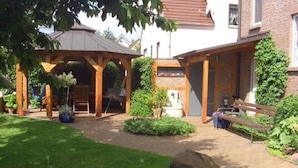 Een tuin met een houten pergola, een bankje en een pad naar een huis.