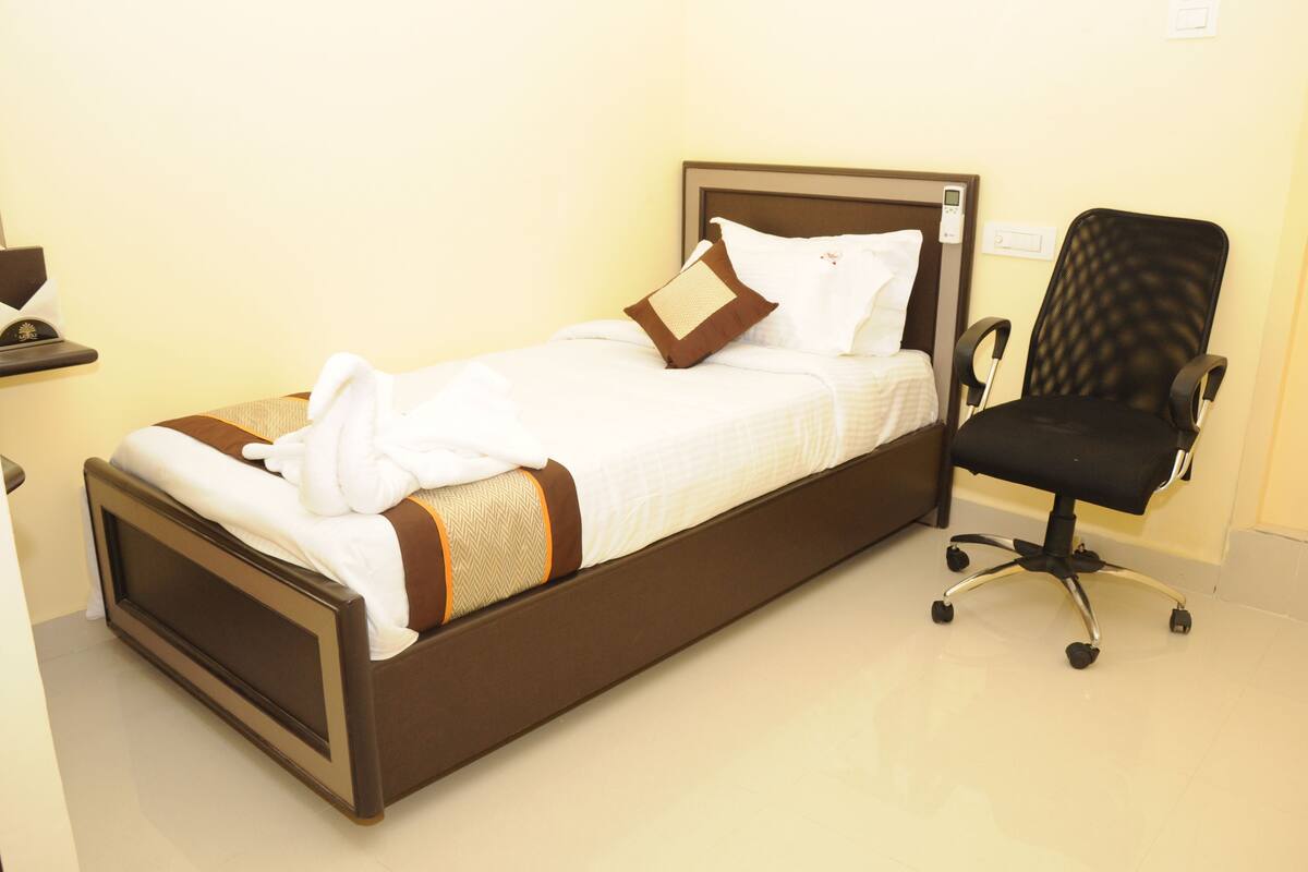 A neatly made bed with a brown headboard, a black office chair, and a small table with a lamp.
