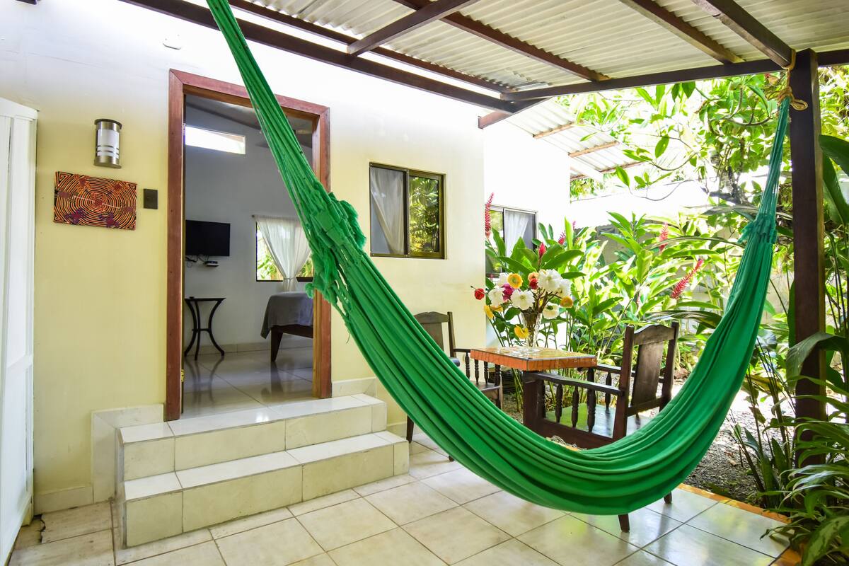 A room with a green hammock, a dining table with chairs, and a staircase leading to another room.