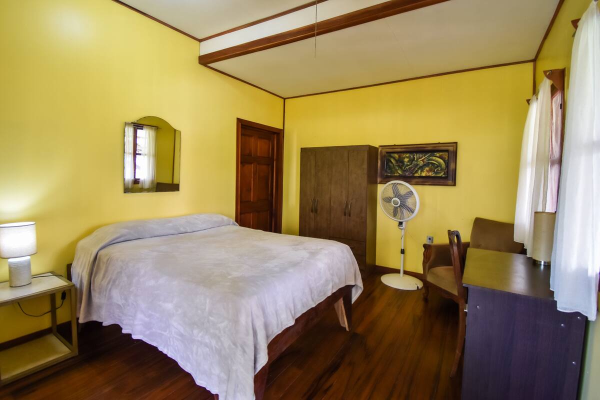 A bedroom with a bed, a nightstand, a wardrobe, a fan, and a desk with a chair.