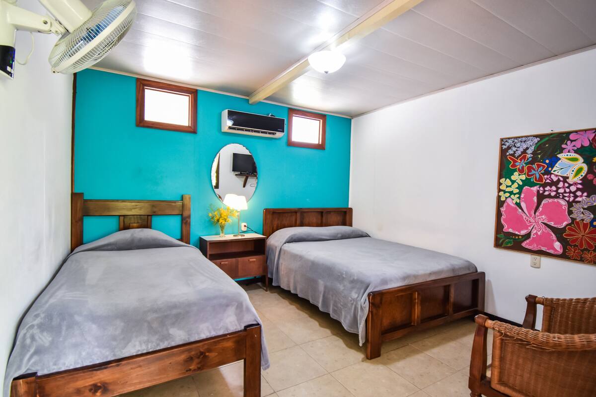 A room with two wooden beds, a fan, a mirror, an air conditioner, and a colorful floral painting on the wall.