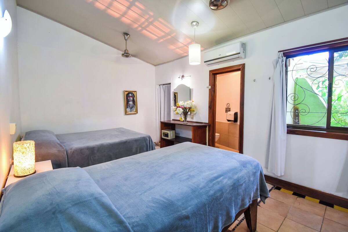 A room with two beds, a bedside table with a lamp, a mirror, a small shelf with flowers, and a window with a view of greenery.