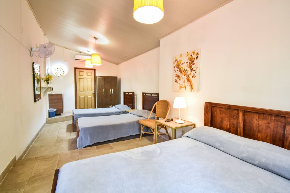 A hotel room with three single beds, a wooden headboard, a small table with a lamp, a chair, and a wall-mounted artwork.