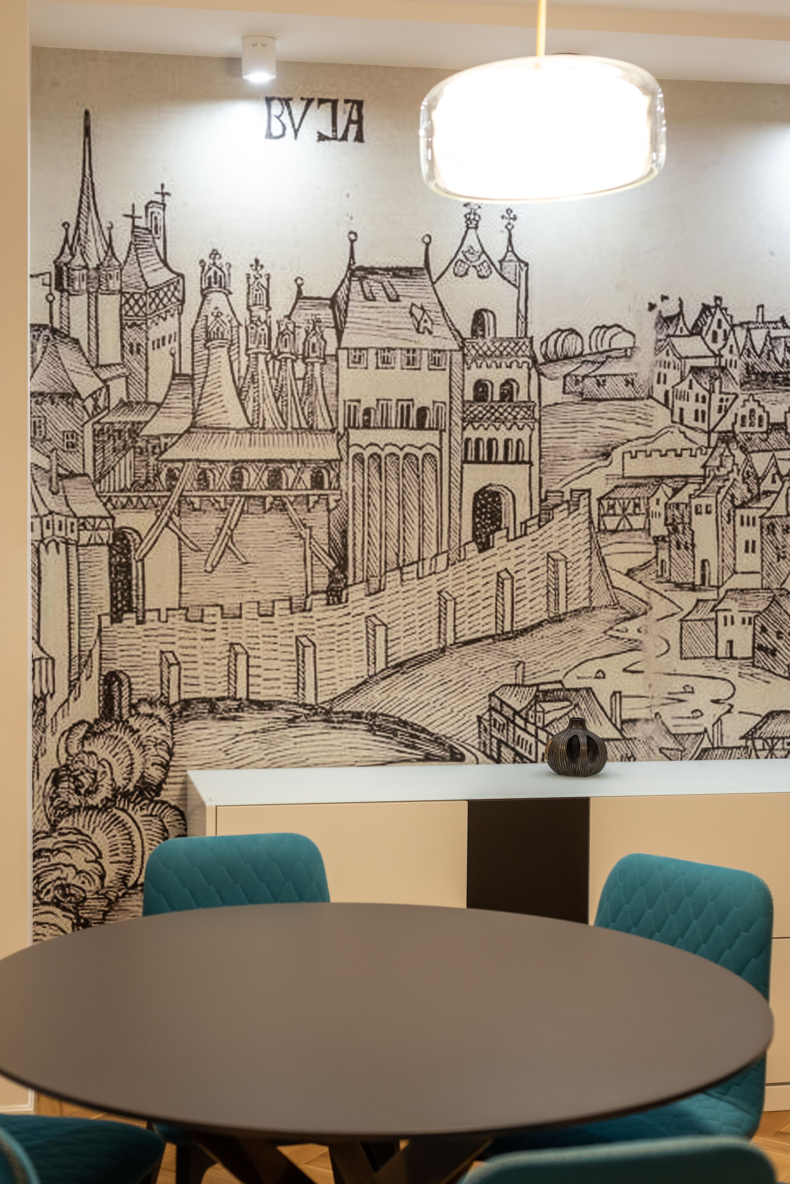 A room with a large wall mural of a medieval town, a round table, and teal chairs.