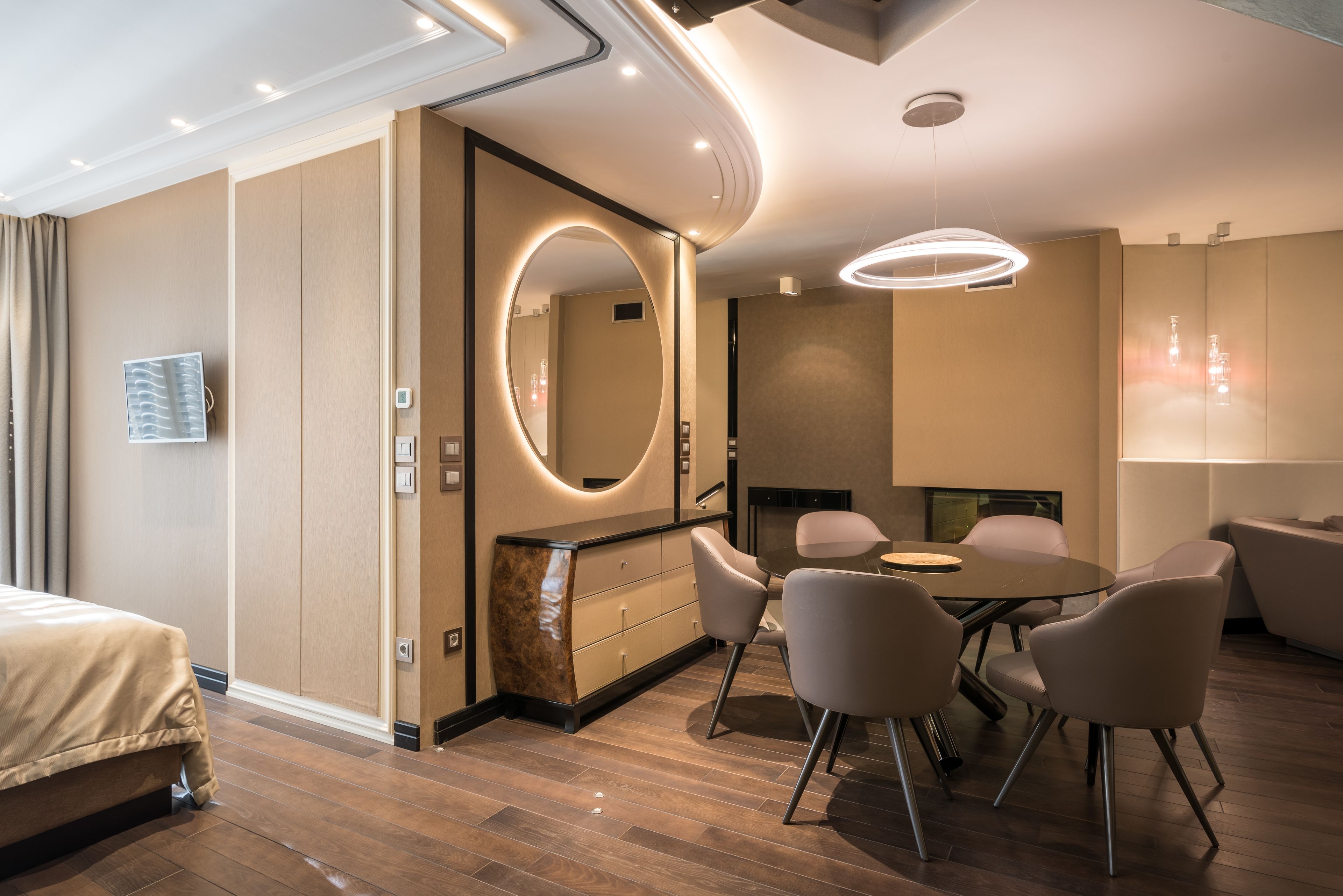 A modern hotel room with a bed, a round mirror, a dining table with chairs, and a fireplace.