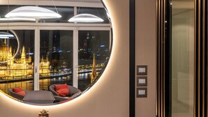 A modern hotel room with a curved seating area, a glass table with a circular light, and a view of a cityscape with a lit-up building. - Castle Apartments Budapest (Budapest)