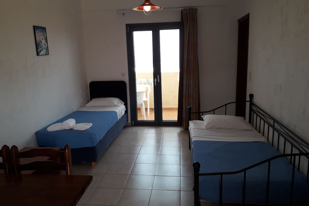 A room with two single beds, a wooden chair, a small table, and a sliding glass door leading to an outdoor area.