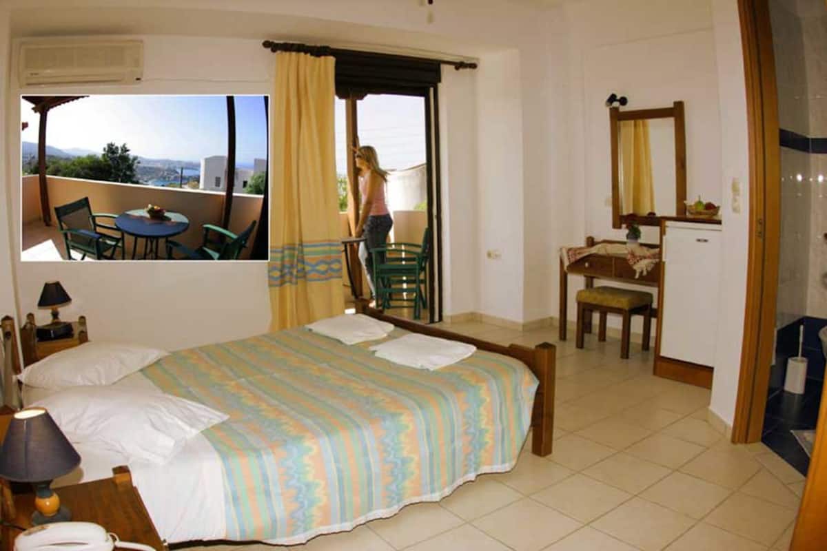 A bedroom with a bed, a desk, a chair, and a small table. There is a balcony with a view of the city.