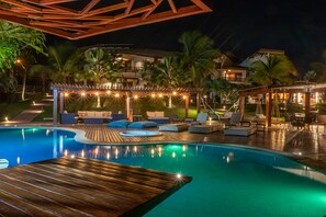 A nighttime pool area with wooden decking, lounge chairs, and a covered seating area with string lights. - Eco Village Cumbuco - Casa Compartilhada (Caucaia)