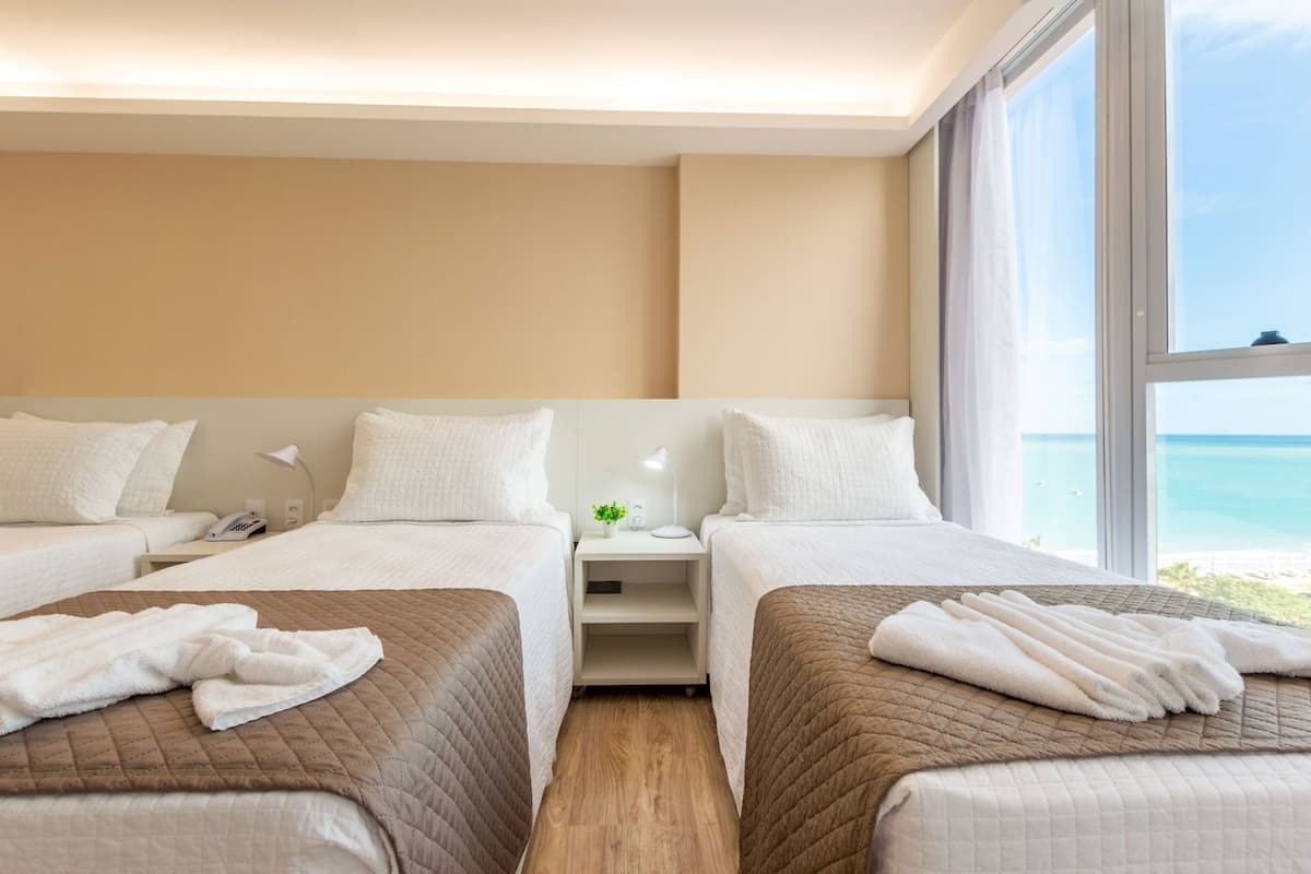 Ein Hotelzimmer mit zwei Betten, einem Nachttisch mit Lampe, einer kleinen Pflanze und Blick aufs Meer.