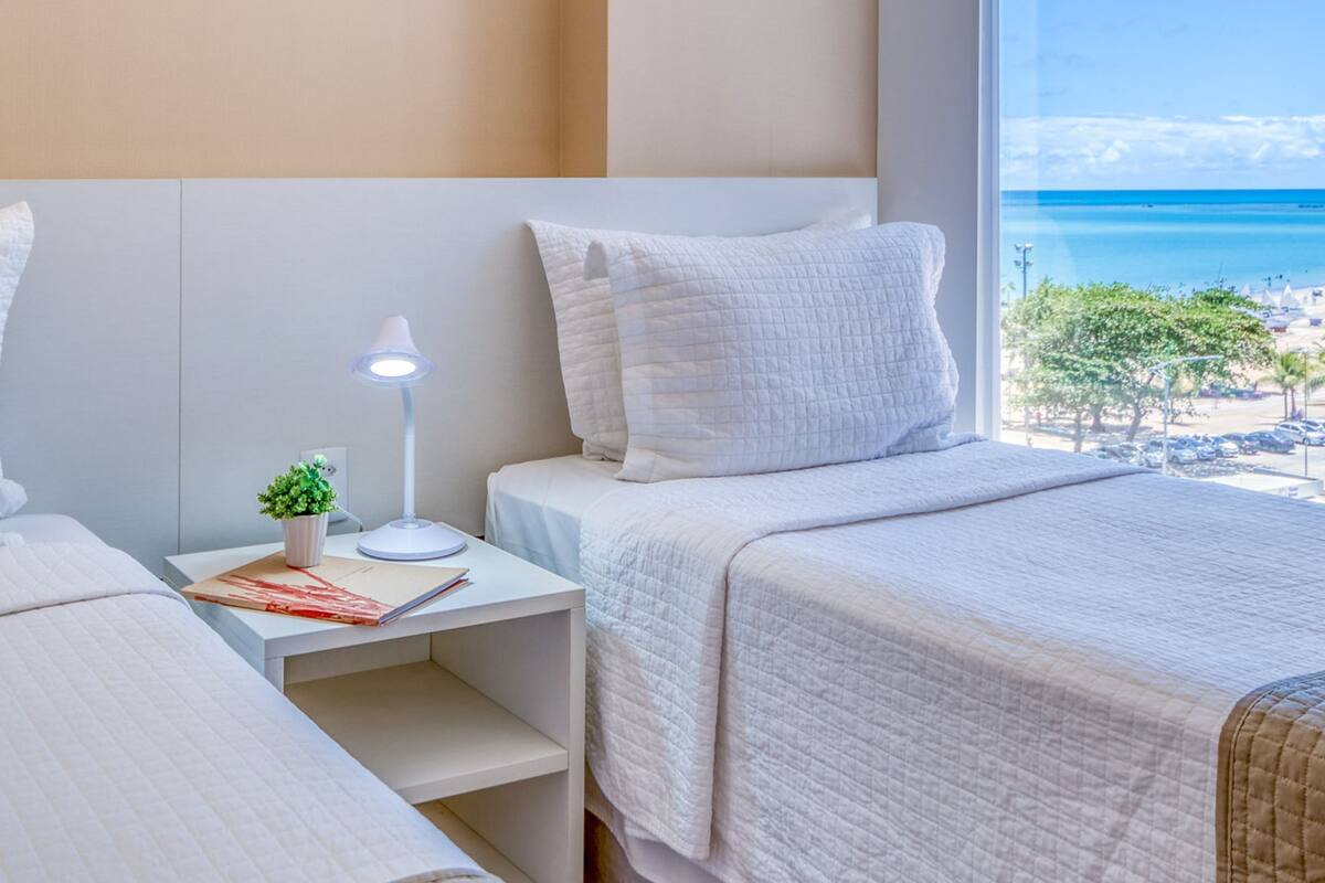 Ein Hotelzimmer mit Bett, Schreibtisch mit Lampe, einer kleinen Pflanze und Blick auf den Strand.