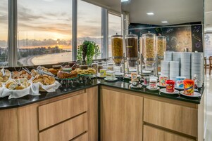 Un bufé de desayuno con diversas opciones de comida y salsas, vista a la ciudad y al atardecer a través de amplias ventanas, y una pared decorativa con una pizarra.