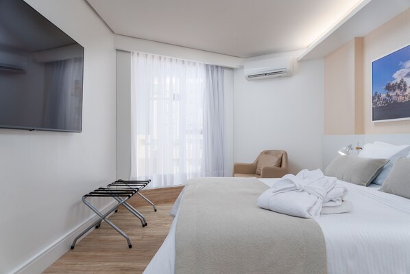 Quarto de hotel moderno com uma cama grande, televisão de tela plana na parede, janela com cortinas leves, uma cadeira e ar-condicionado fixado na parede.