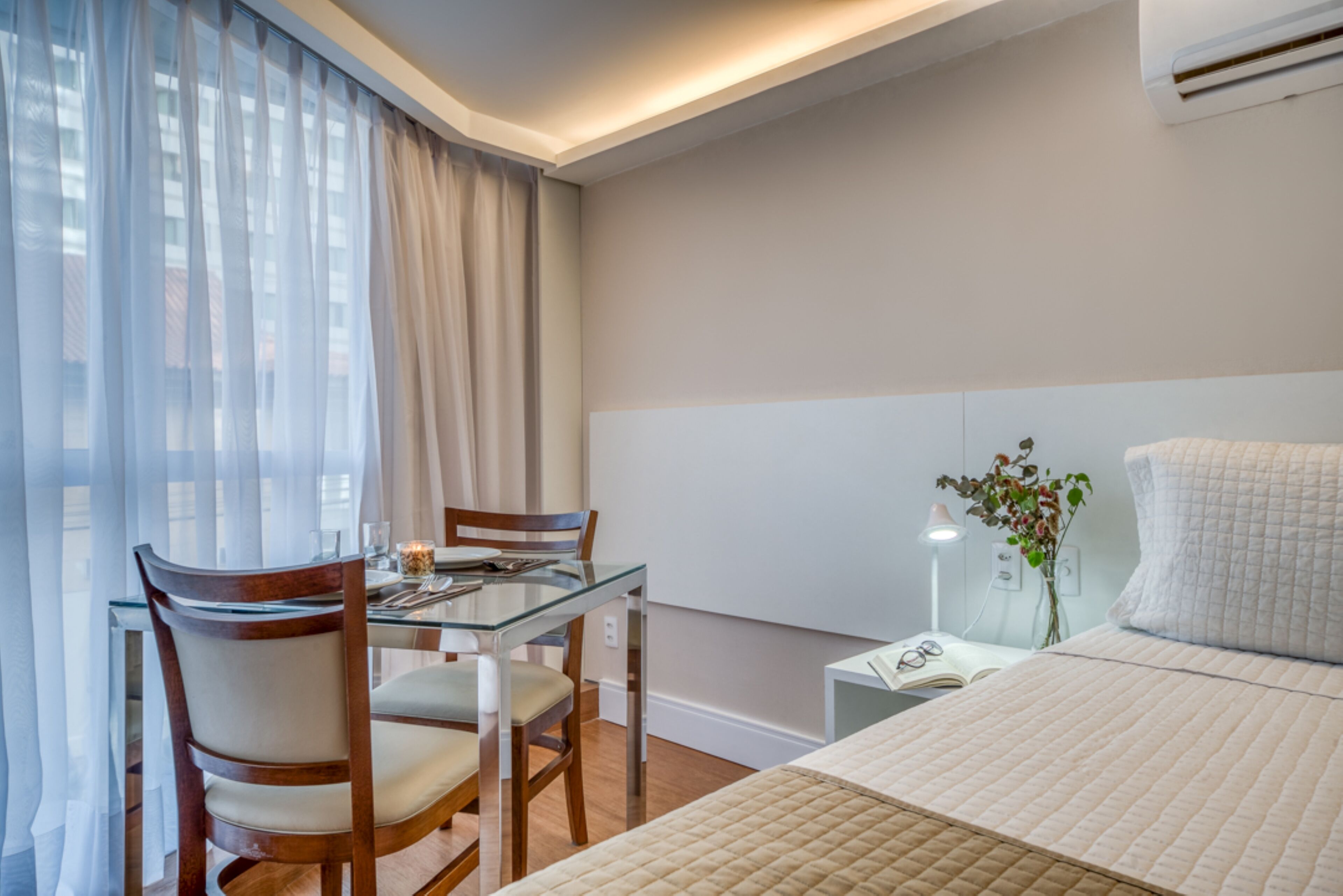 A hotel room with a glass dining table, two chairs, a bed with a quilted cover, a bedside table with a lamp, and a window with sheer curtains.