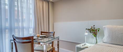 A hotel room with a glass dining table, two chairs, a bed with a quilted cover, a bedside table with a lamp, and a window with sheer curtains.