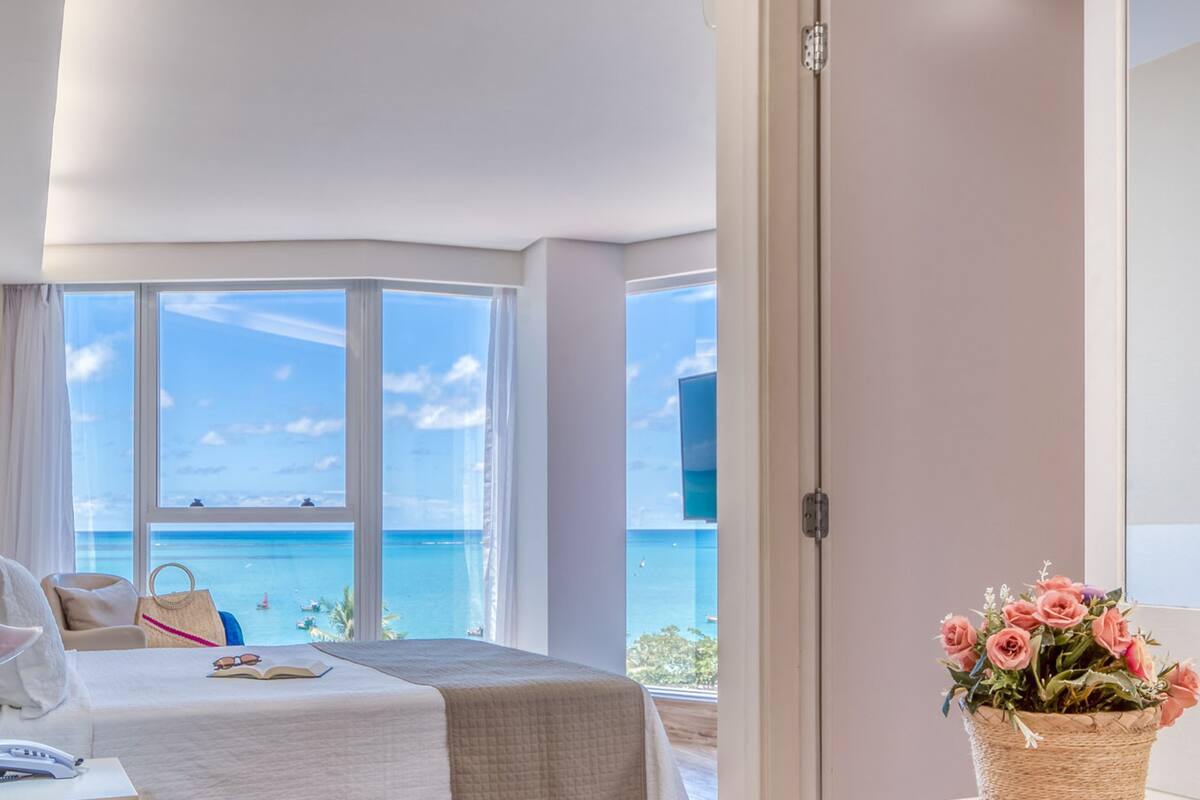 Ein modernes Hotelzimmer mit einem großen Fenster und Meerblick. Es gibt ein Bett, einen Schreibtisch und einen Stuhl.