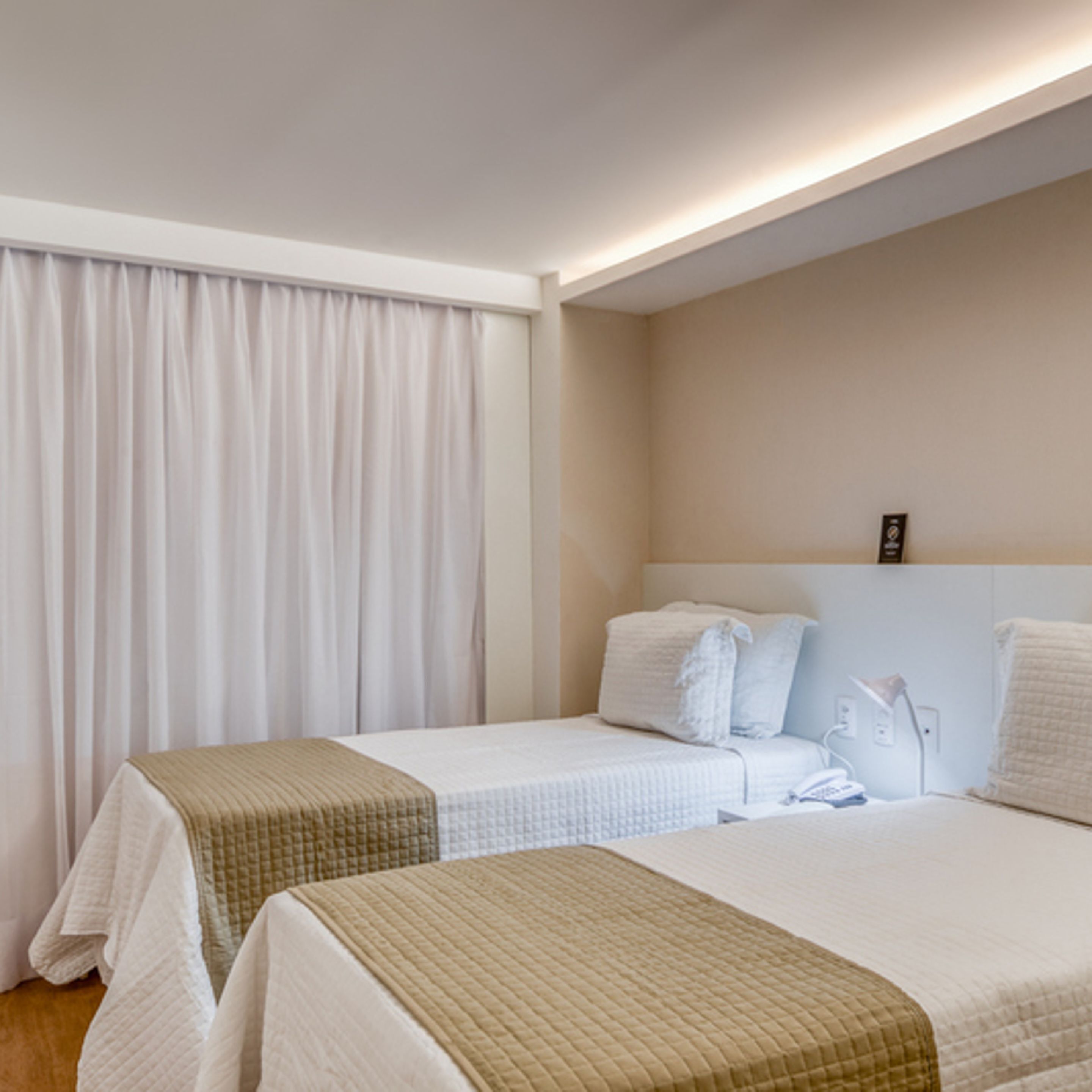 A hotel room with two beds, a large window with curtains, a bedside table with a lamp, and a wall-mounted phone.