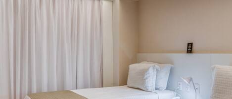 A hotel room with two beds, a large window with curtains, a bedside table with a lamp, and a wall-mounted phone.