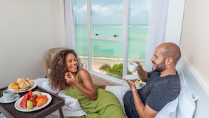 A couple sitting on a bed with a breakfast tray, a cup, and a plate of fruit. - Hotel Brisa Suítes Pajuçara (Maceió)